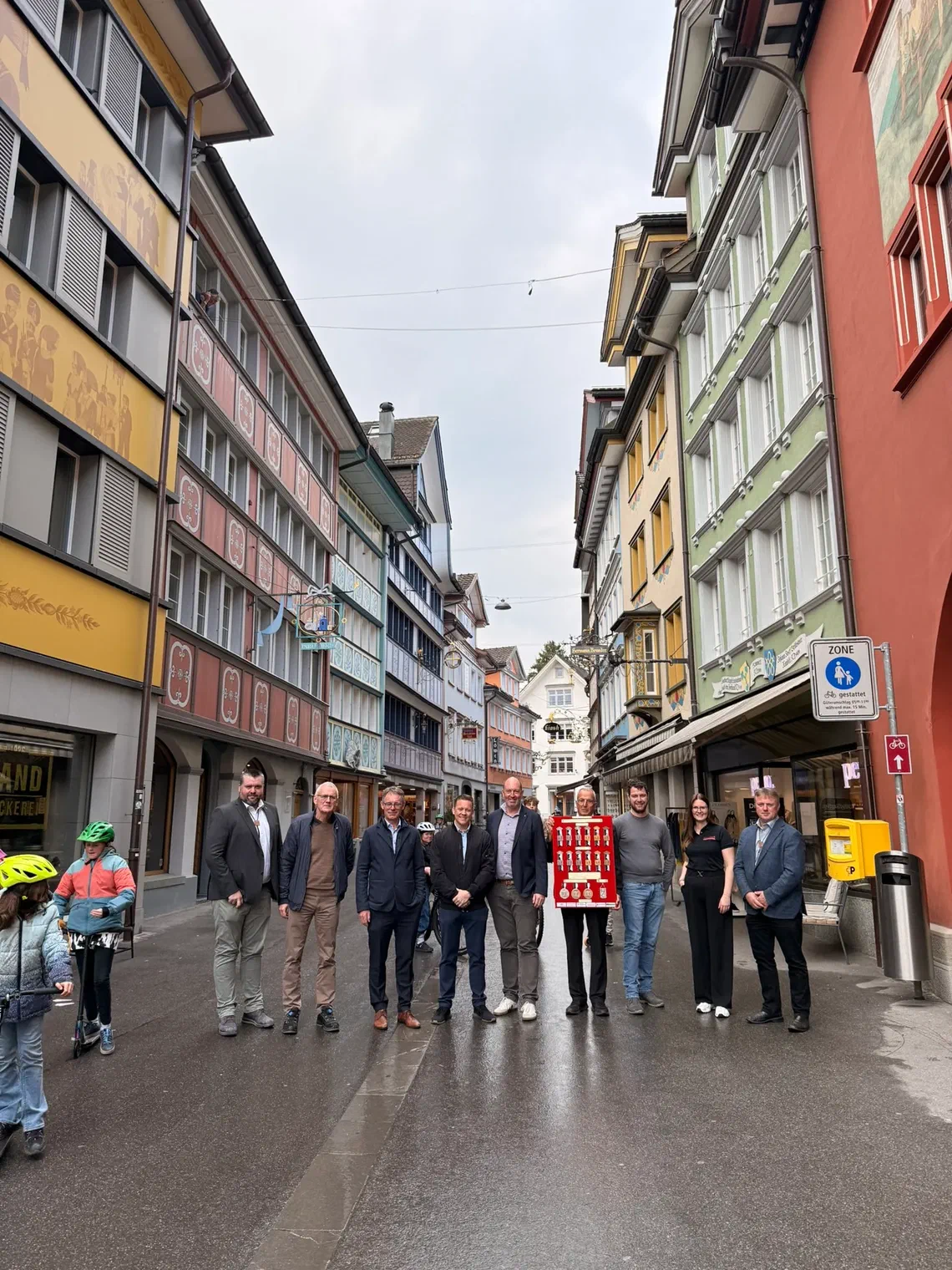 Neun Personen stehen in der Altstadt von Appenzell vor bemalten Häuserfassaden, einer hält eine rote Tafel mit der Kranzserie des AIKSF 2028.