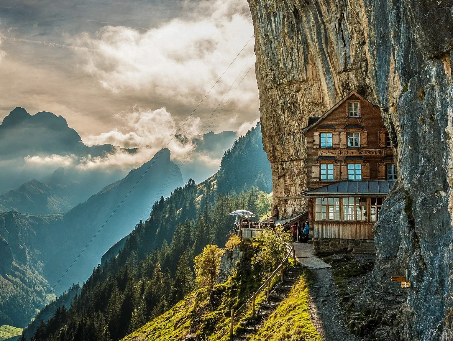 Berggasthaus Äscher unter der überhängenden Felswand der Ebenalp, davor Gäste auf der Terrasse, dahinter der Alpstein in Wolken.