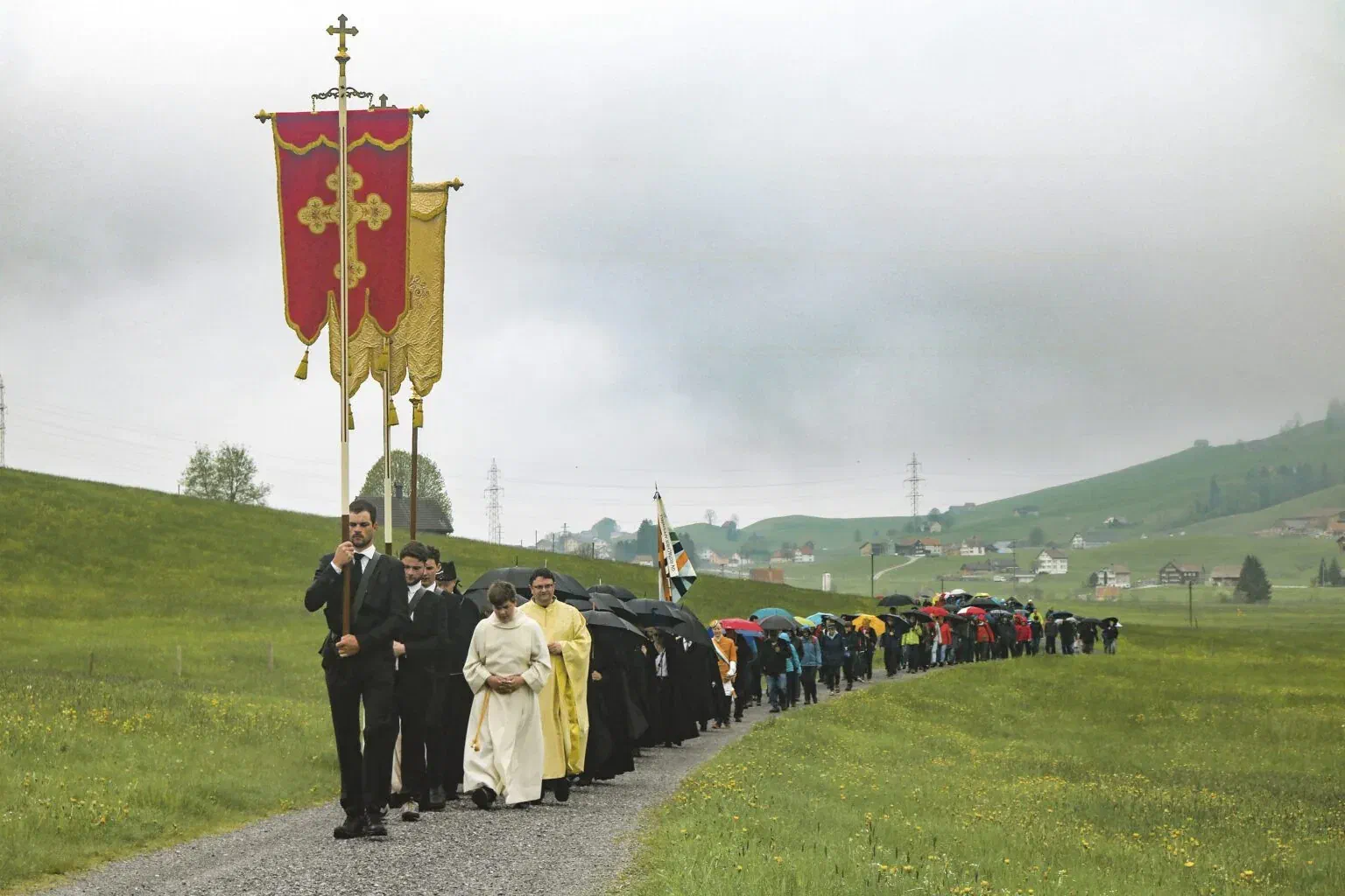 Prozession mit Kreuz und Fahnen zieht bei Regen über die Wiesen zum Stoss.